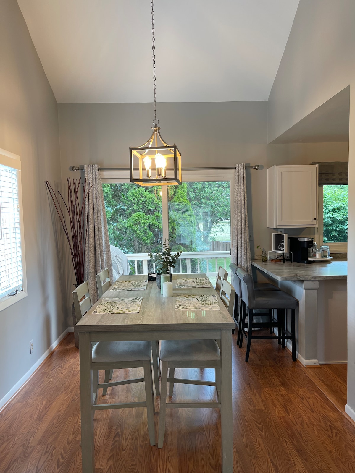 A dining area features a rustic wooden table surrounded by six light-colored chairs. Natural light floods in through a large window, highlighting the muted gray walls. A pendant light hangs above, and a small coffee station is visible in the corner of the adjoining kitchen.