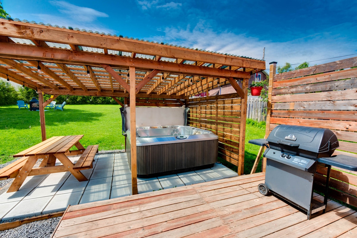An outdoor area features a covered hot tub next to a barbecue grill and a picnic table. The space is surrounded by greenery, providing an inviting atmosphere for relaxation and gatherings. Paved flooring complements the wooden deck, enhancing usability.