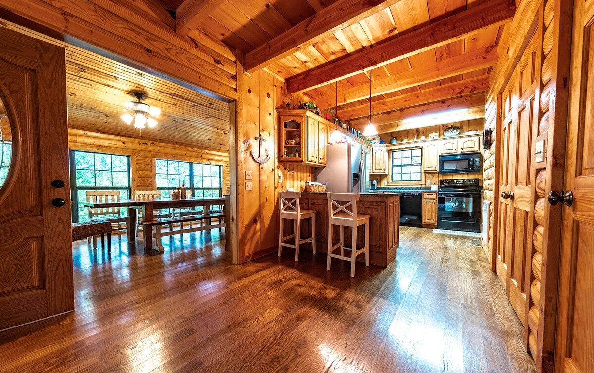 The cabin's interior showcases warm wooden paneling and an open floor plan. A spacious kitchen features wooden cabinets and modern appliances, with seating at a bar counter. A dining area with a large table is visible through an archway, accompanied by plentiful natural light from large windows.