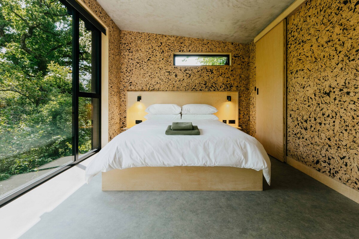An open bedroom features a spacious bed dressed in white linens atop a light wood frame. Natural light floods the room through large windows, showcasing the surrounding greenery. Textured walls add a unique element to the minimalist design, while practical bedside lighting provides gentle illumination.