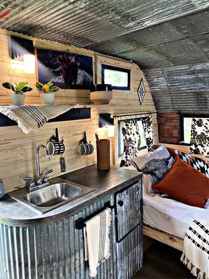 An interior space features a rustic design with corrugated metal walls and wooden accents. A compact kitchen area is visible, equipped with a sink and several hanging mugs. A cozy sleeping area includes patterned bedding and decorative pillows, enhancing the inviting atmosphere of the camper.
