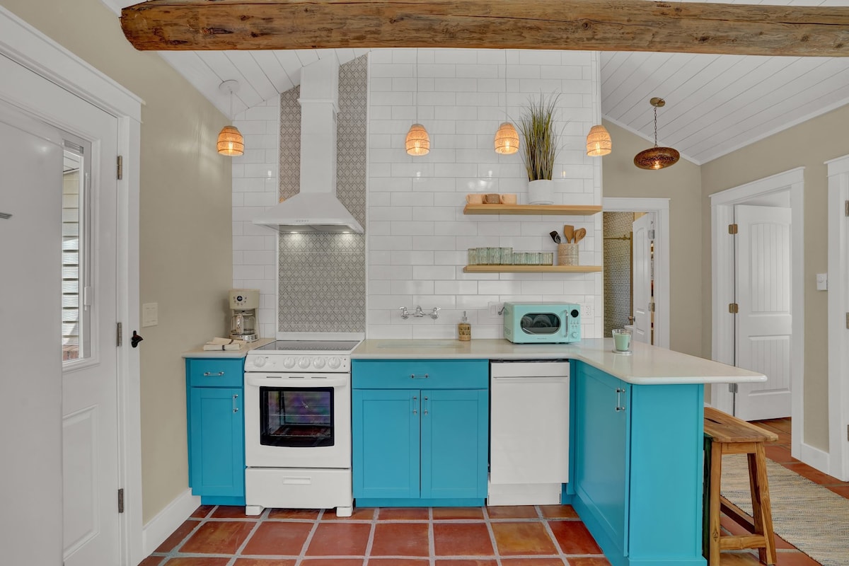 A bright kitchen features turquoise cabinetry paired with white countertops. Modern appliances, including a stove and microwave, are visible. Pendant lights hang from the ceiling, adding warmth to the space. A textured backsplash creates visual interest, and open shelving displays tasteful kitchenware.