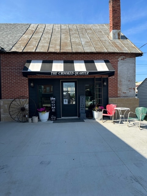 The exterior of The Crooked Quarter features a welcoming entrance with a striped awning and flower pots. Two small tables with chairs are placed outside, inviting guests to relax. The brick façade and roof details highlight a charming small-town atmosphere.