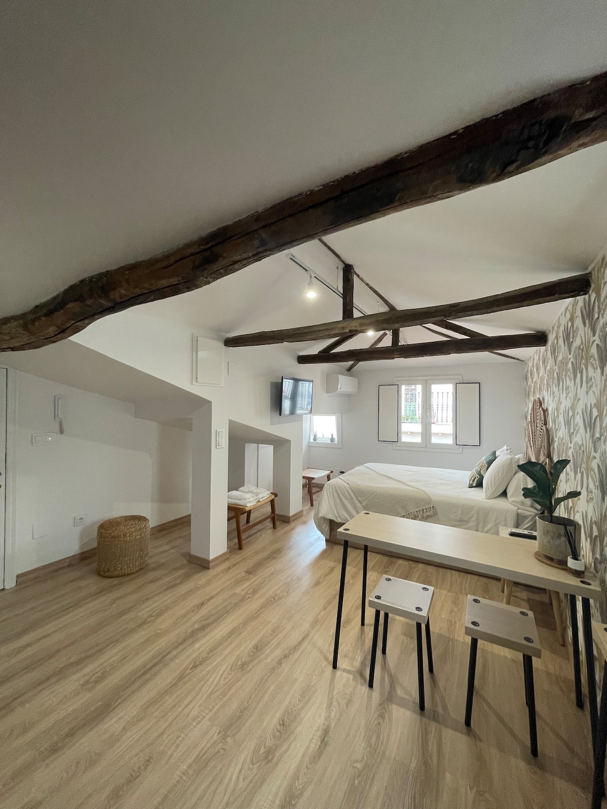 An open and airy attic space is highlighted by original wooden beams. A spacious bed is accompanied by a small dining area featuring two stools and a table. Natural light filters through windows, illuminating the light wooden flooring and a decorative wall with a subtle pattern.
