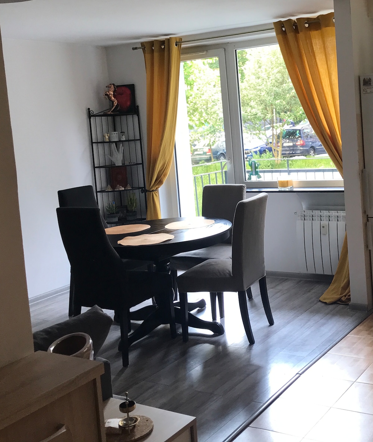 A dining area features a round table surrounded by four chairs, two in dark tones and two in light colors. Tall windows with golden curtains allow natural light to fill the space, while decorative items and plants can be seen on a nearby shelf.