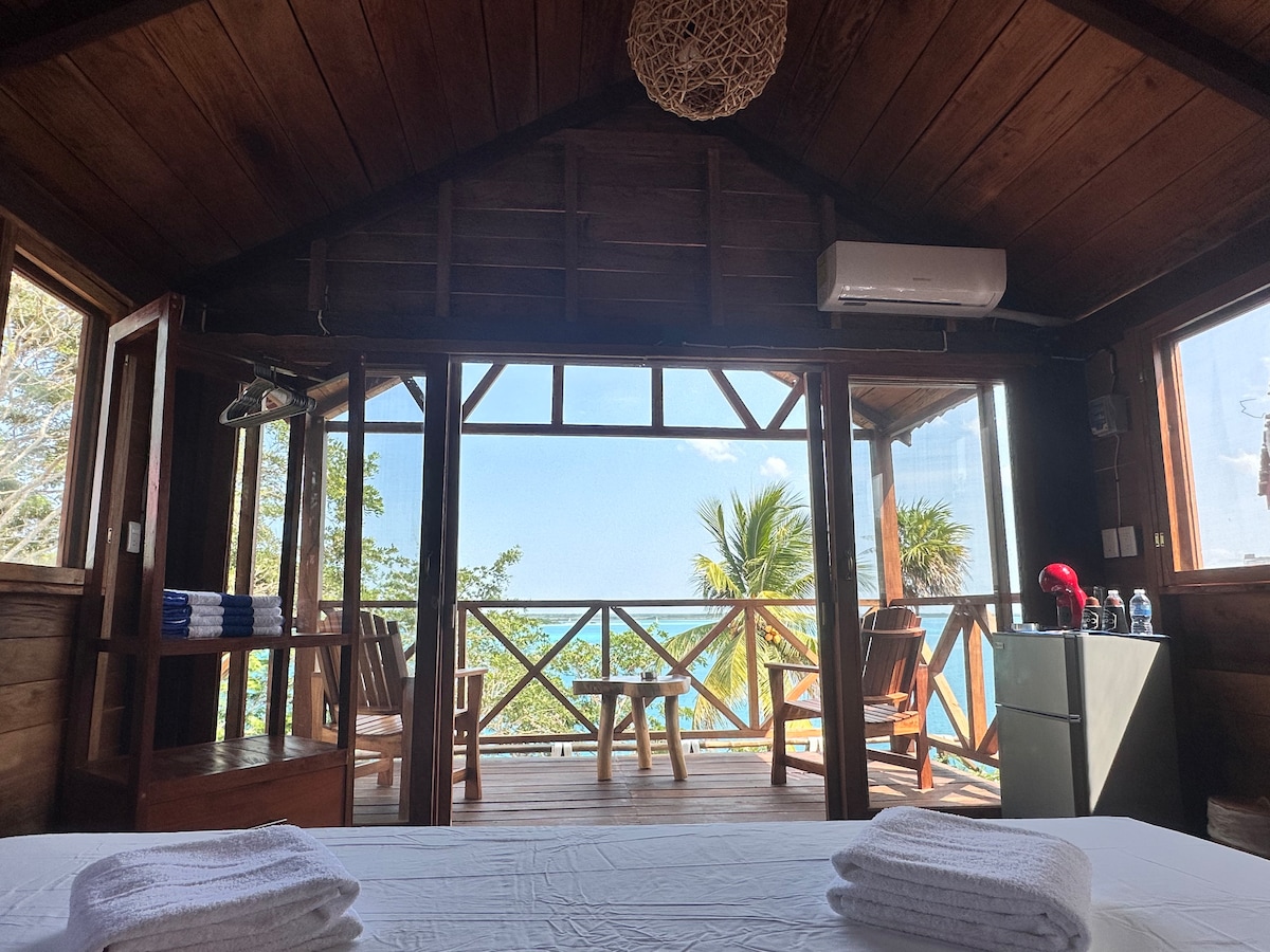 A wooden cabin interior features large glass doors that open to a private terrace with seating. The space includes a spacious bed with clean linens and towels, while warm wooden accents and natural light enhance the serene atmosphere.