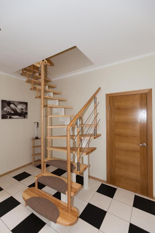 A spiral staircase made of wood and metal is seen, leading to an upper level. The surrounding area features a light-colored wall and a door with a wood finish. The floor is tiled in a checkerboard pattern of black and white, complemented by a simple shelf and wall art.