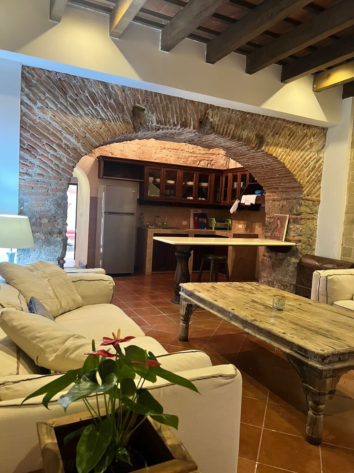 A cozy living area is framed by an arched brick wall, featuring a soft seating arrangement with light-colored sofas. A rustic wooden coffee table is placed in the center. In the background, the kitchen area is visible, showcasing dark wood cabinetry and modern appliances.