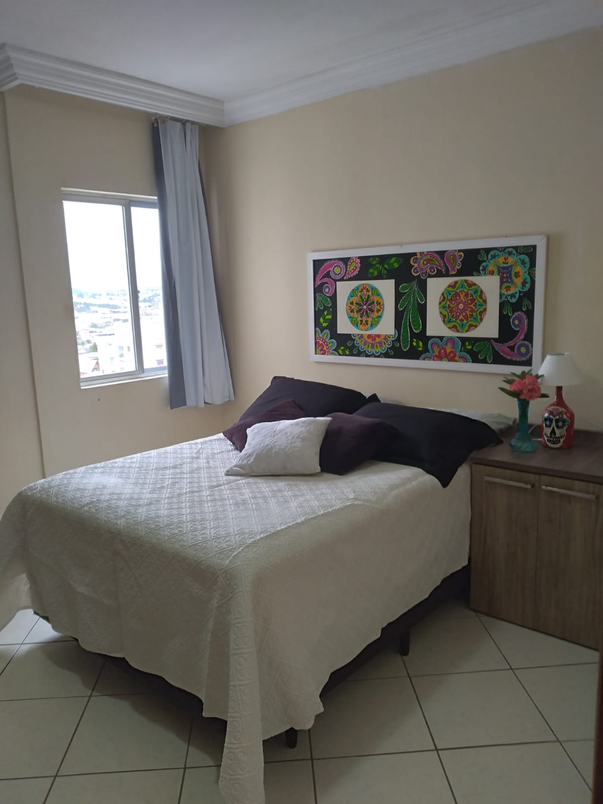 The sleeping area features a spacious bed with a white quilt, complemented by dark pillows. A colorful wall art piece enhances the ambiance. Nearby, a small bedside table holds a decorative skull and a lamp, while a window allows natural light to fill the room.