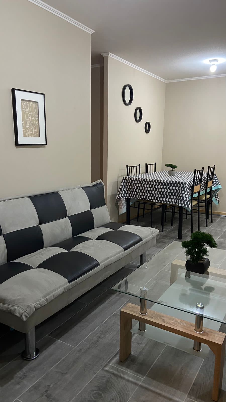 A living area features a stylish grey and black sofa alongside a glass coffee table. A dining space is visible in the background, furnished with a patterned table and black chairs. Decorative wall art enhances the neutral-toned walls.
