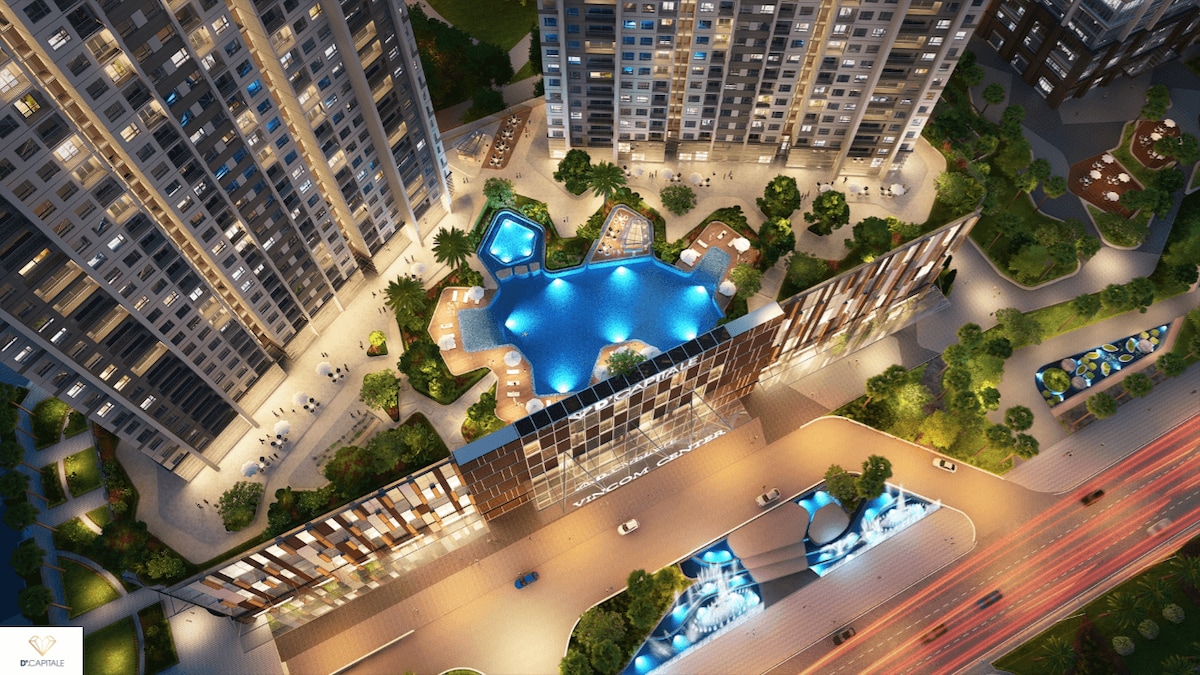 An aerial view of an urban complex showcases a vibrant outdoor swimming pool bordered by carefully landscaped greenery. The building's modern architecture features illuminated balconies and an inviting entrance, with vehicles visible on the road below, contributing to a lively atmosphere.