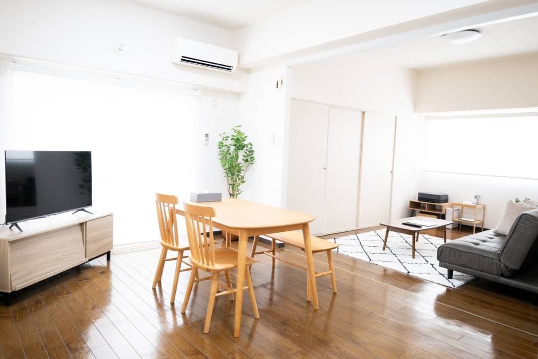 A bright and open living area features a wooden dining table surrounded by four chairs. A flat-screen TV is positioned on a media console. A sofa bed and a small coffee table are placed across from the dining space, with large windows providing ample natural light.