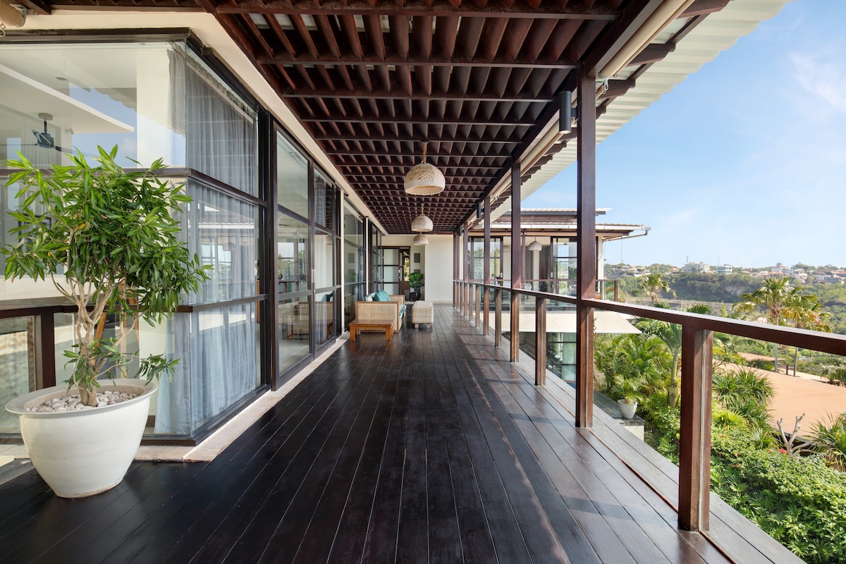 A spacious balcony is lined with dark wooden flooring and surrounded by glass railings. Potted plants are placed along the edges, and natural light brightens the area. The view extends to the lush greenery and the property’s landscaped surroundings.