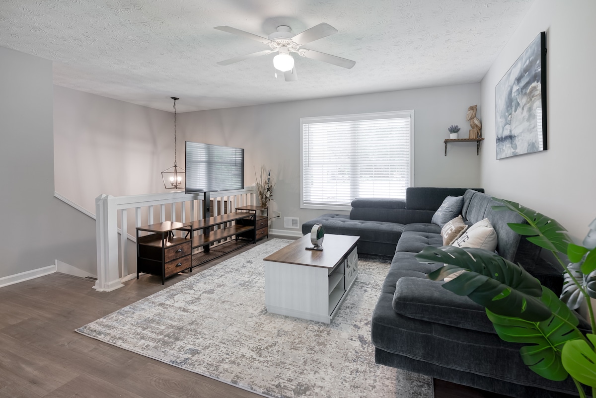 A spacious living area features a large sectional sofa and a wooden coffee table. Natural light fills the room through a wide window, and a neutral-colored area rug anchors the space. A ceiling fan contributes to airflow, and decorative elements are tastefully arranged.