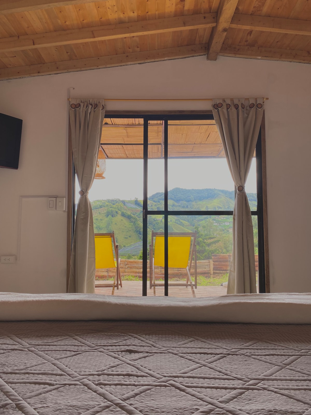 A view from the interior showcases a large glass door leading to a balcony with two bright yellow chairs. The room features a textured bedspread, and the landscape outside displays rolling green hills under a clear sky.