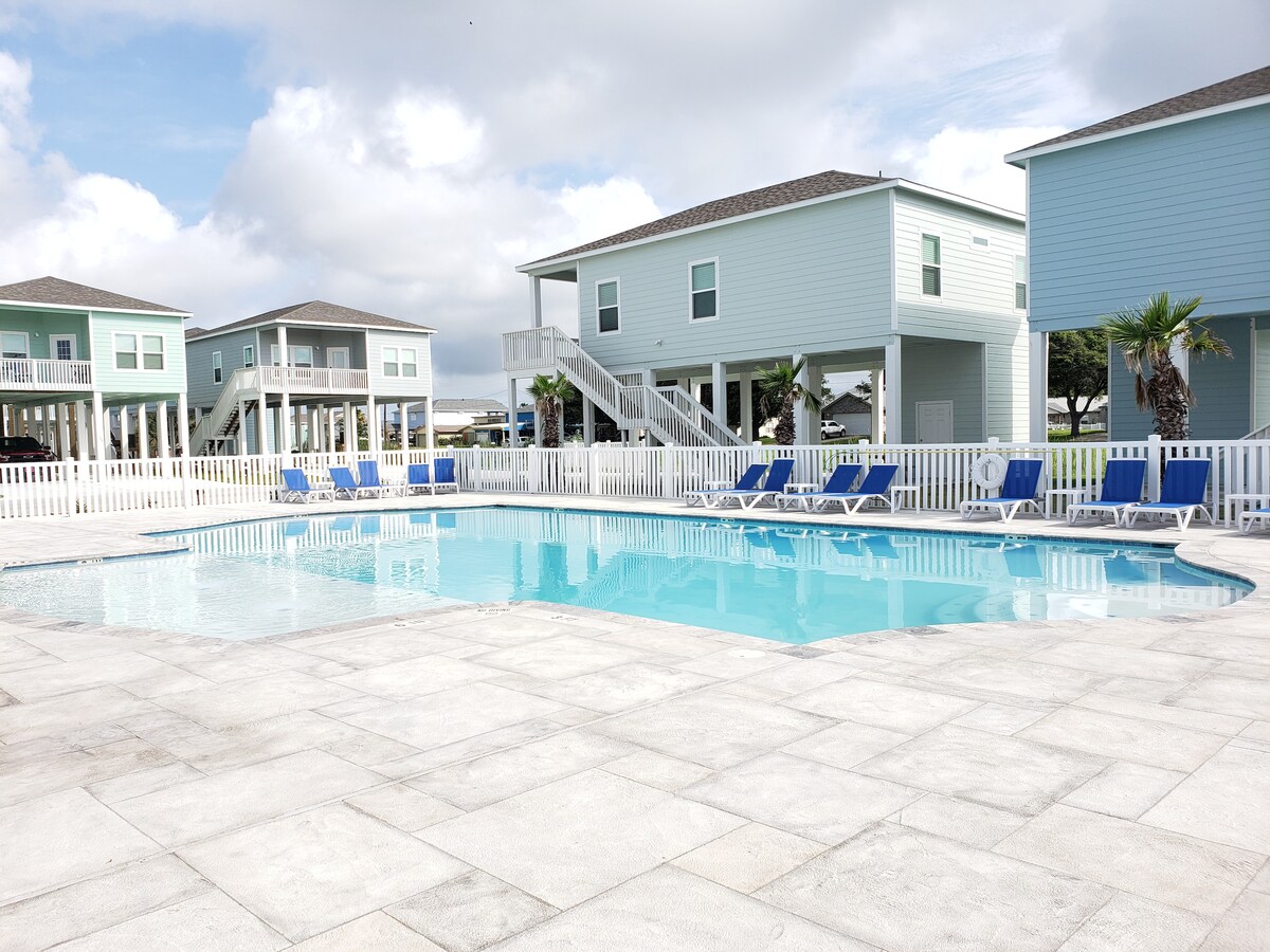 A clear view of a swimming pool surrounded by a white fence, with several blue lounge chairs positioned along the edges. The light-colored tiled area creates an open space, while several beach-style homes are visible in the background against a partly cloudy sky.