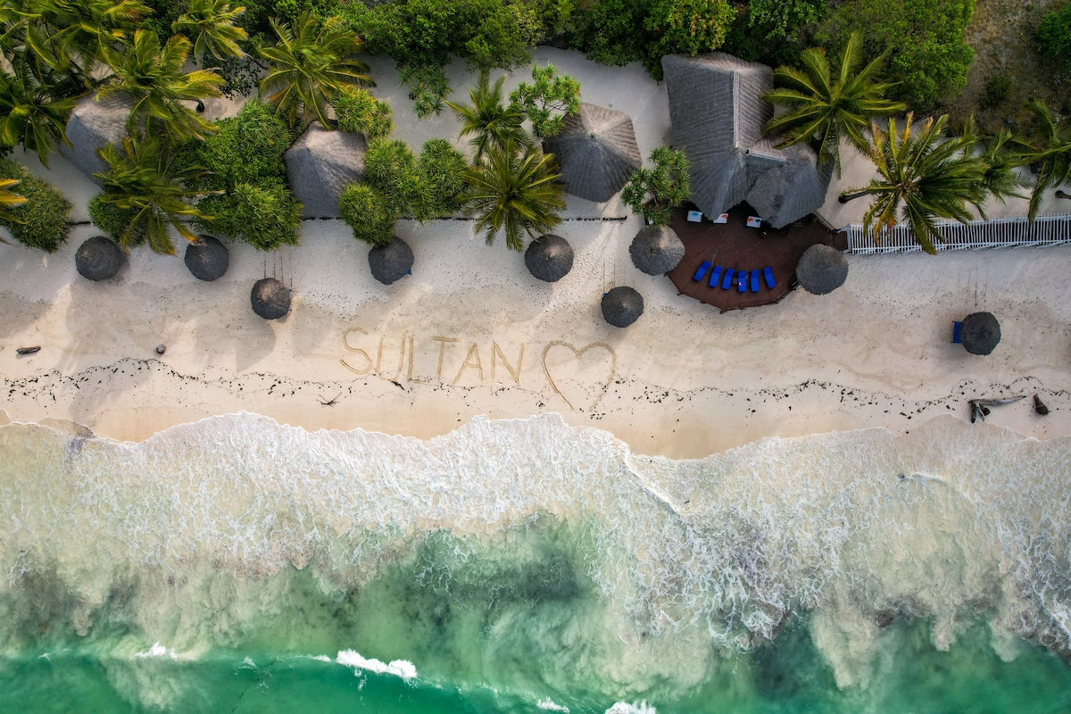 An aerial view captures a stretch of sandy beach lined with several thatched umbrellas and loungers. The clear, emerald water gently laps at the shore, where the word 'SULTAN' is written in the sand, surrounded by a heart shape.