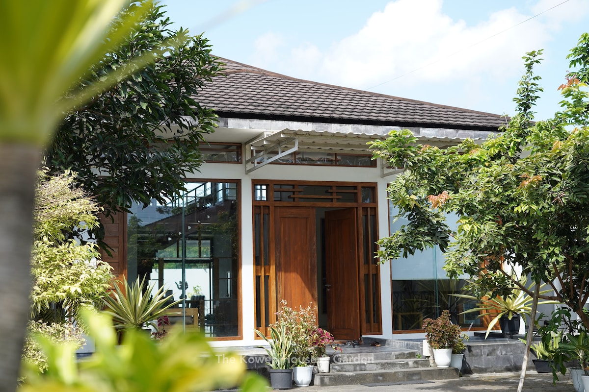 The entrance is framed by lush greenery, featuring a spacious patio with various potted plants. Large sliding glass doors allow natural light to enter, while a traditional roof design complements the modern aesthetic of the house.