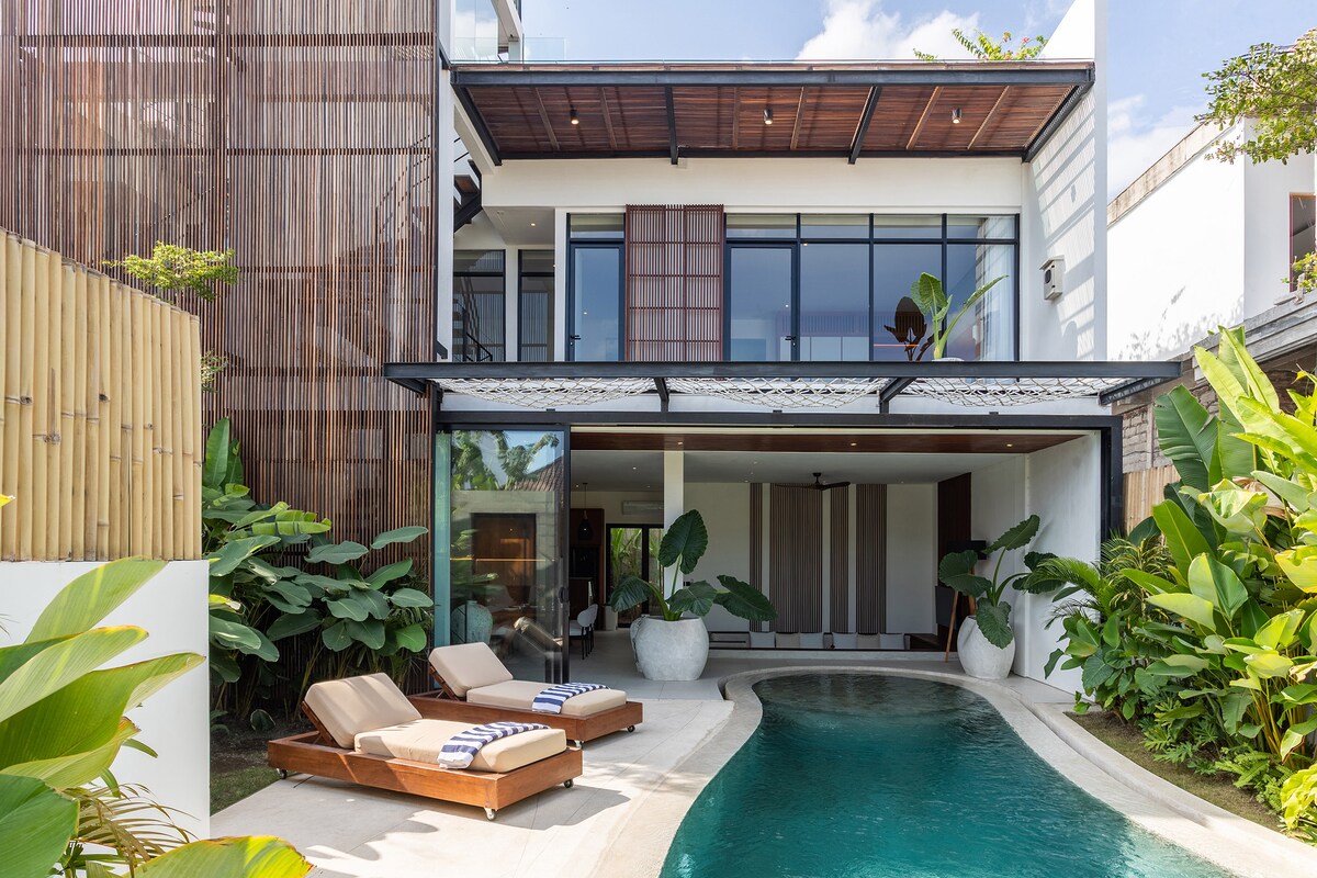 A modern villa exterior is surrounded by lush greenery, featuring a refreshing plunge pool. Elegant sun loungers are placed alongside the water. Expansive glass doors open to the interior, allowing natural light to flow into the living space.