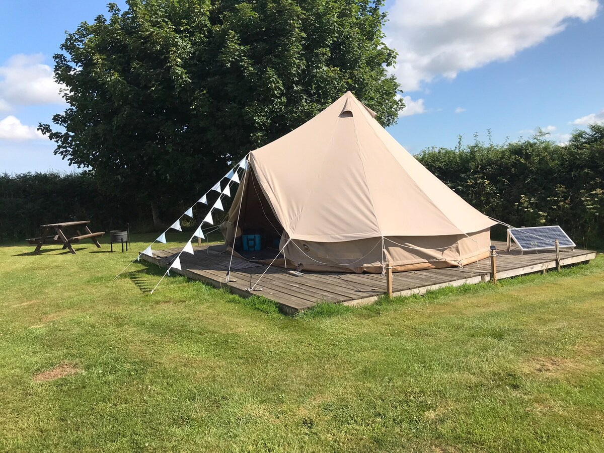 A spacious bell tent is situated on a wooden platform surrounded by a well-kept grassy area. Solar panels are mounted on the tent, and decorative bunting hangs along the entrance, offering a welcoming touch. A picnic table is nearby, providing an outdoor dining option.
