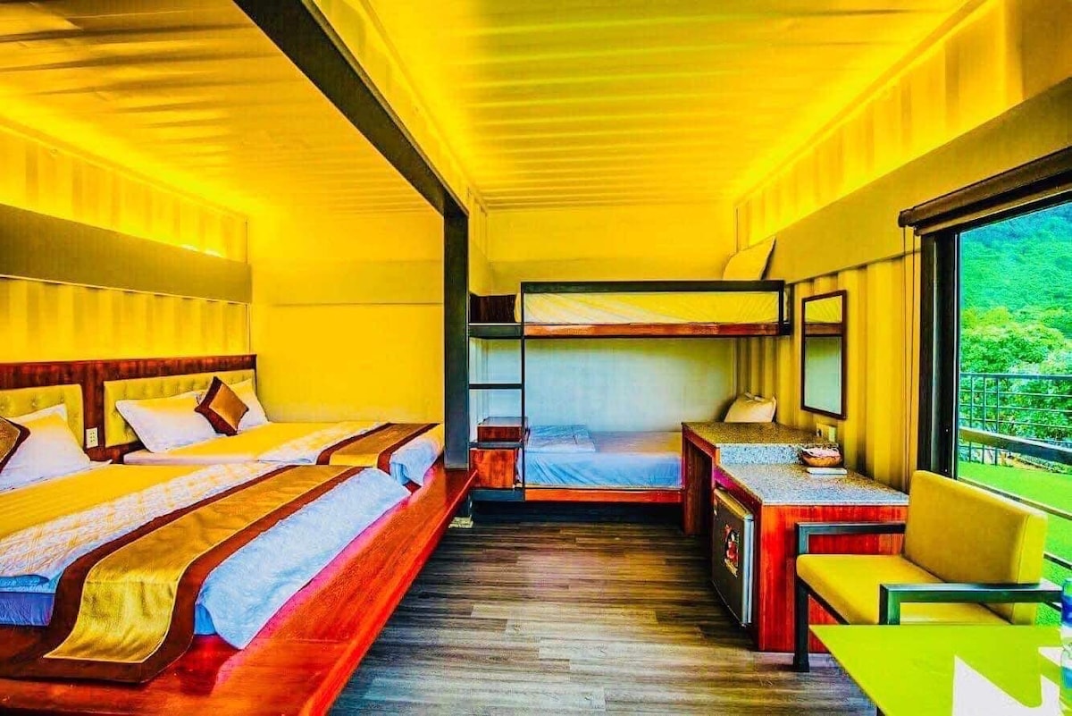 A bright and spacious room features two large beds with crisp linens, complemented by a cozy bunk bed. A desk and chair are positioned near a window, allowing natural light to enhance the warm tones of the walls and furnishings.