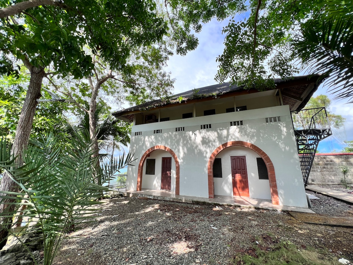 A two-story building is shown, featuring an exterior with arched windows framed in red brick. The structure is surrounded by lush greenery, with tall trees and tropical plants. A metal staircase is visible on one side, leading to the upper level.
