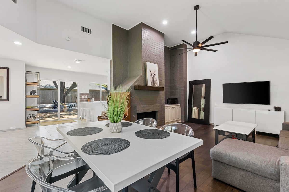 A bright living area features an open layout with a modern fireplace and a large flat-screen television mounted on the wall. A rectangular dining table is set with placemats, surrounded by transparent chairs. Natural light enters through expansive windows, illuminating the space.