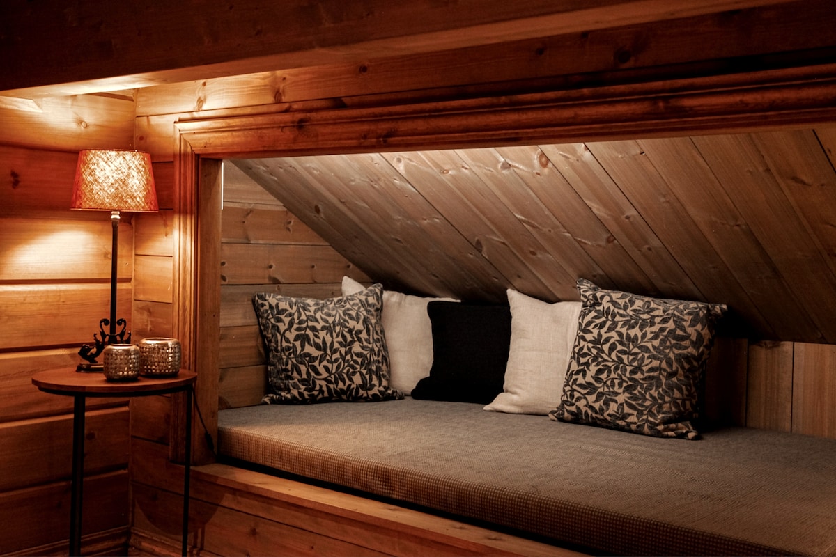 A cozy nook is created with a comfortable mattress surrounded by wooden walls. Soft cushions in varying patterns are arranged for relaxation. A small side table holds decorative items and a warmly lit lamp, adding to the inviting atmosphere.