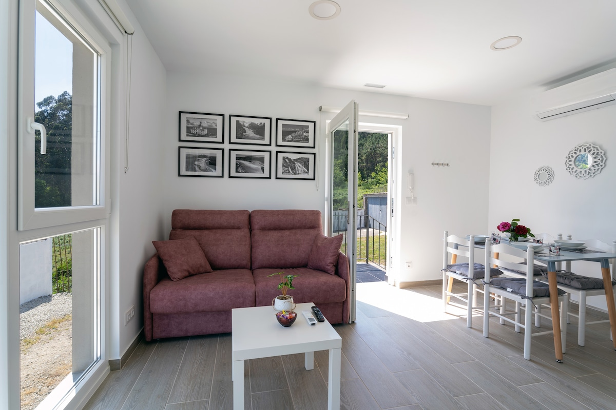 A light-filled living area features a comfortable pink sofa and a coffee table. Black and white photographs adorn the wall above the sofa. Large glass doors open to the outdoor space, while a dining table with white chairs is visible in the background.