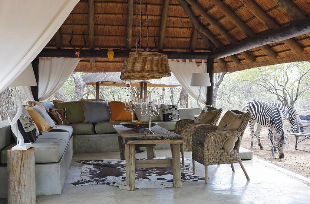 An open-air lounge area features a thatched roof with comfortable seating arranged around a rustic wooden table. Neutral-colored cushions and a decorative rug add warmth to the space. Natural light filters through sheer drapes, while a zebra can be seen nearby in the surrounding bush.