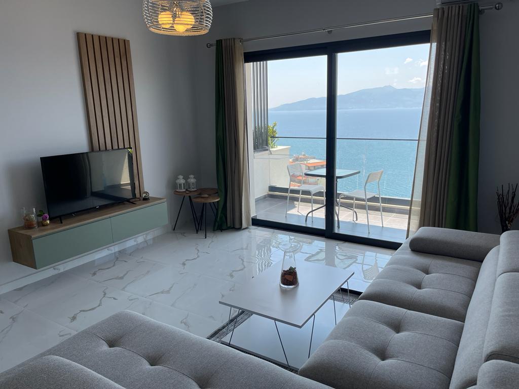 A modern living area features large glass sliding doors that open to a stunning sea view. A sleek gray sofa is positioned near a contemporary coffee table, while a television sits on a minimalist cabinet. Light-colored curtains frame the windows, enhancing the bright environment.