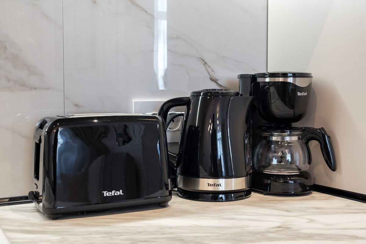 A modern kitchen countertop showcases a collection of black Tefal appliances, including a toaster, electric kettle, and coffee maker. The appliances are neatly arranged against a textured white marble background, creating a stylish and functional cooking space.
