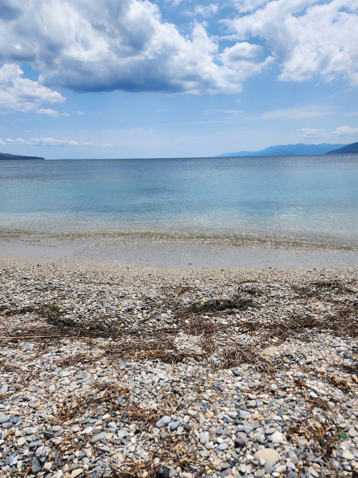 A calm beach setting is captured, featuring smooth pebbles along the shore with scattered seaweed. Gentle waves create a serene shoreline, while the clear blue water extends towards distant hills under a partly cloudy sky.