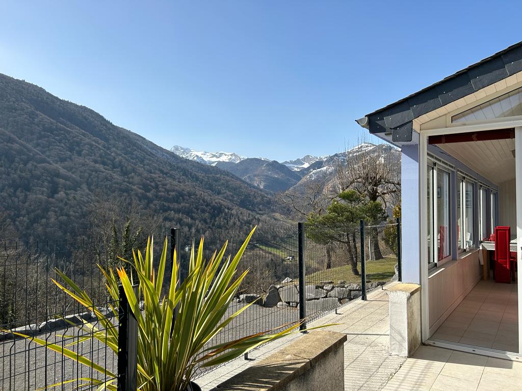 A spacious outdoor terrace is visible, bordered by a low fence. Mountain ranges stretch into the distance, capped with snow under a clear blue sky. A potted plant adds a touch of greenery to the scene, which showcases the natural beauty of the surrounding landscape.