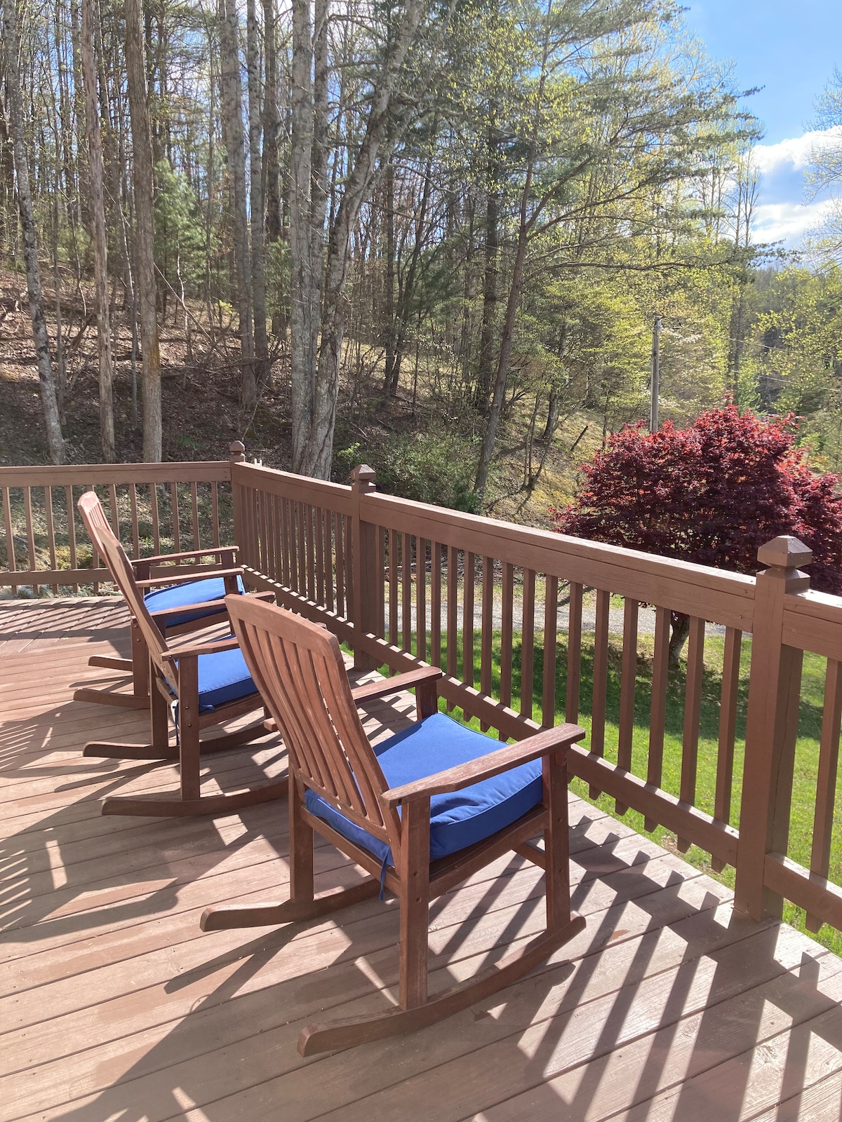 Two wooden rocking chairs with blue cushions are placed on a spacious deck. The railing provides an open view of the surrounding trees and greenery, creating a serene outdoor space ideal for relaxation.