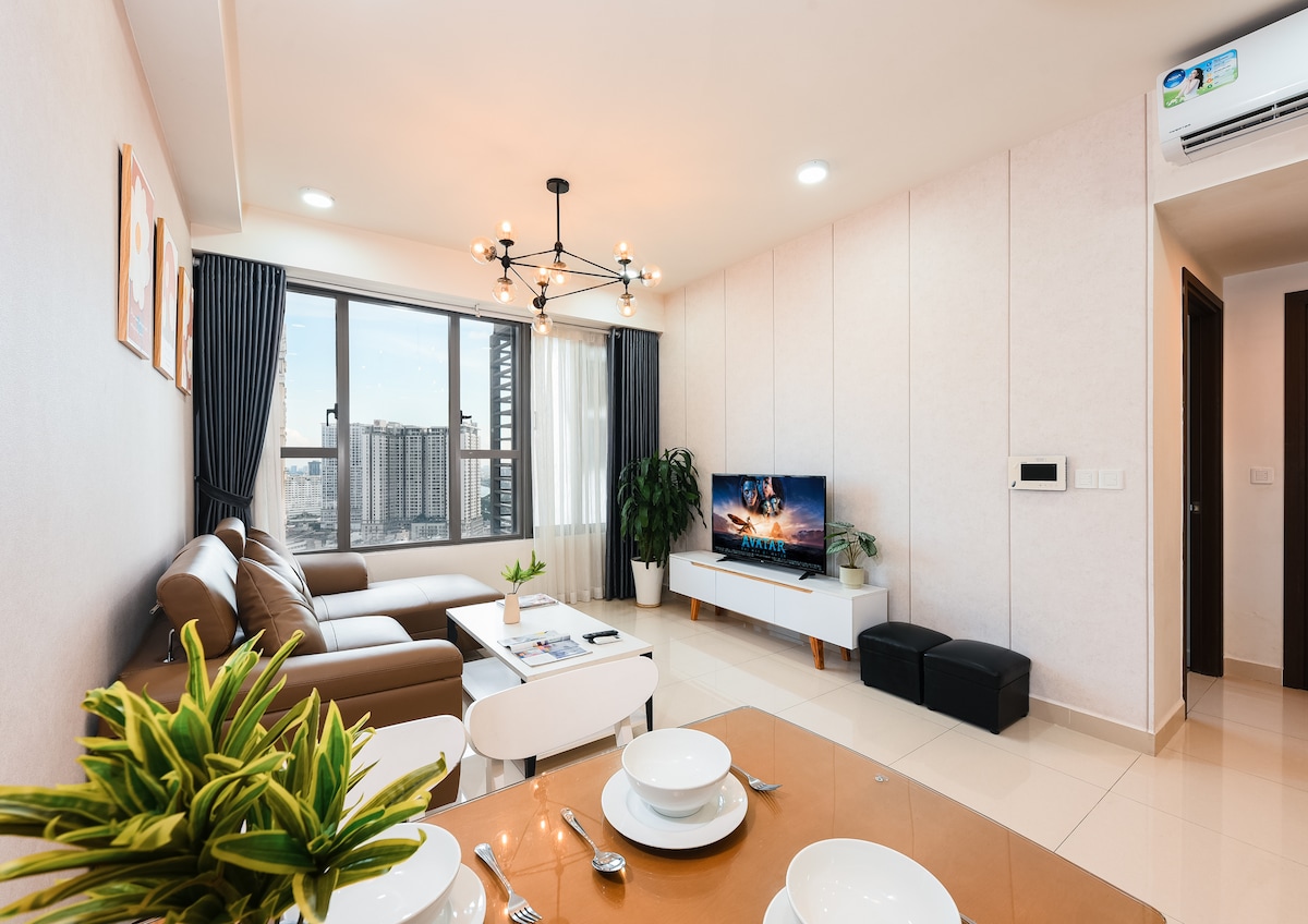 A modern living area is visible, featuring a comfortable brown sofa and a large window allowing natural light. A sleek TV stand displays a flat-screen television. A dining table is set with dishes, while a green plant adds a touch of nature to the space.