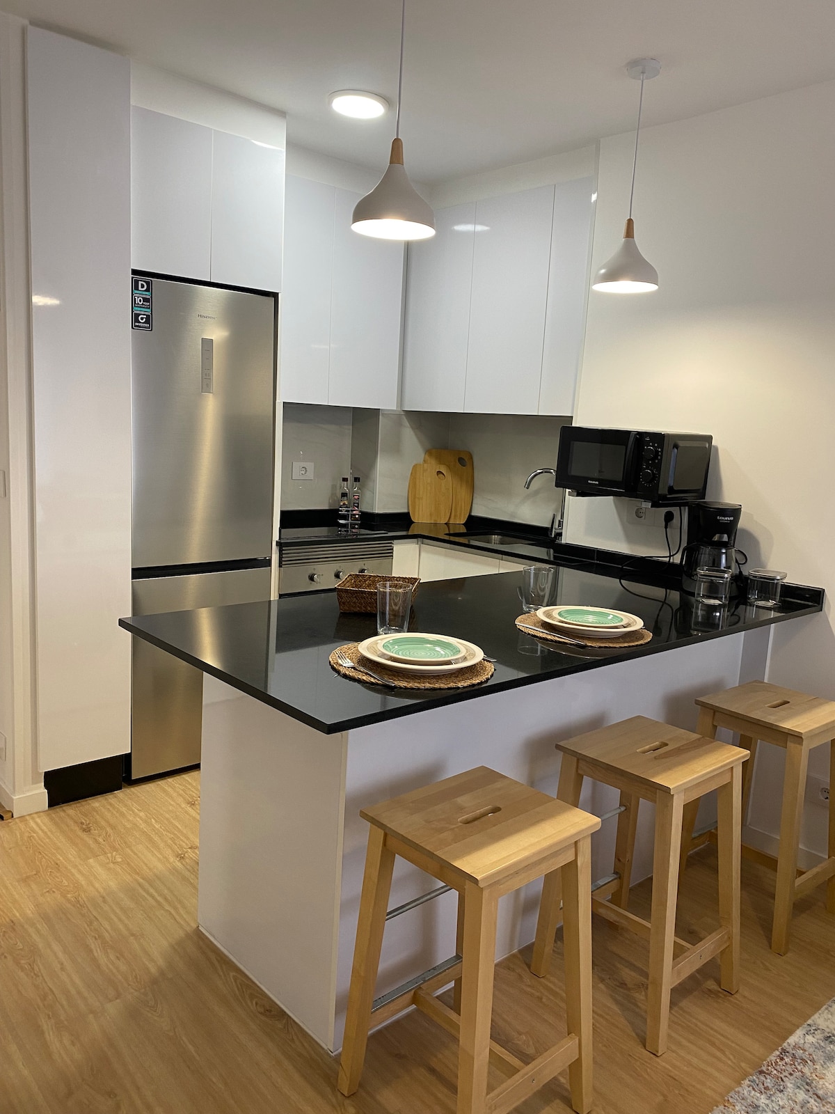 A modern kitchen space features sleek white cabinetry and a black countertop. Wooden bar stools are positioned around a breakfast bar, set with plates and glasses. Stainless steel appliances, including a refrigerator and microwave, are clearly visible against a neutral backdrop.
