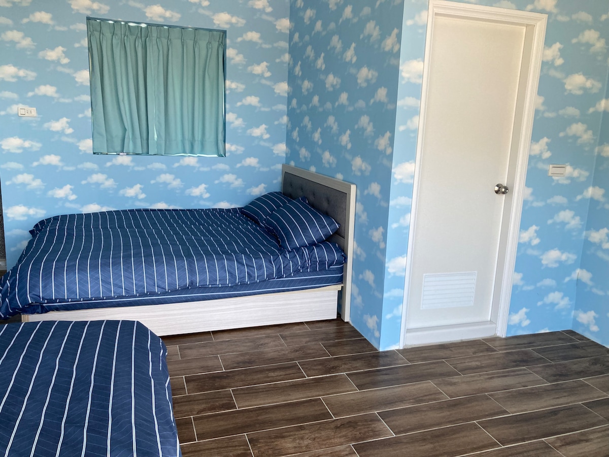 A room featuring two spacious beds with striped blue bedding, set against a whimsical cloud-patterned wall. A window with light blue curtains allows natural light to fill the space. A white door is visible, leading to the hallway.