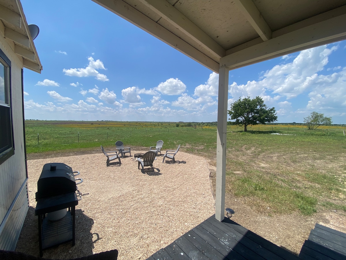 An outdoor area is depicted featuring a gravel circle arranged with several chairs and a fire pit at its center. A grill is positioned nearby under the shade of the porch. A vast view of grassy fields and scattered trees extends into the distance.