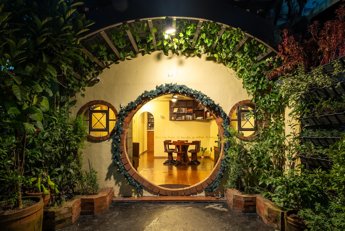The entrance of the Hobbit house features a whimsical round doorway framed by rich greenery. A warm glow from inside highlights a wooden dining table with chairs, inviting guests into a cozy, nature-inspired space. The exterior is adorned with a variety of lush plants.