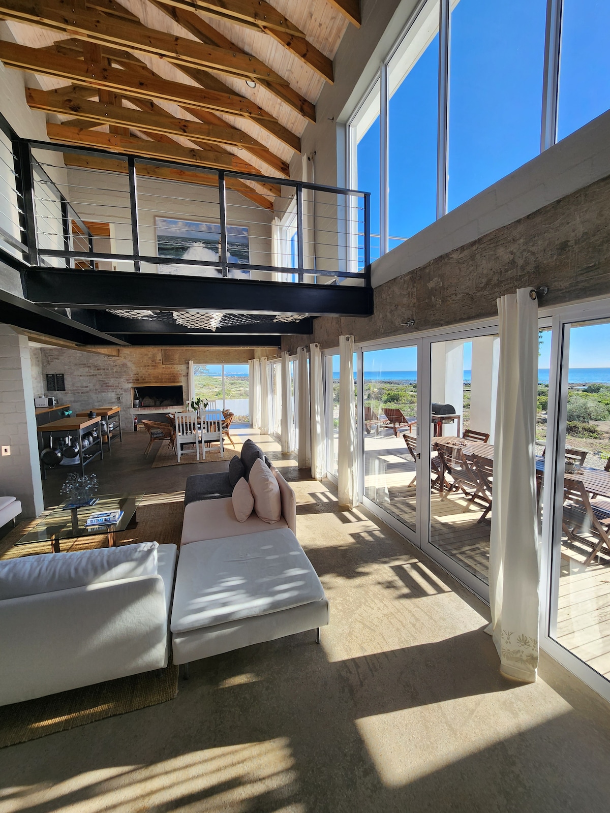 A spacious living area is illuminated by natural light streaming through large glass windows. The high ceilings feature exposed wooden beams. A soft grey sofa is accompanied by light-colored cushions, while a dining table and outdoor seating are visible in the background, with a serene view of the landscape.