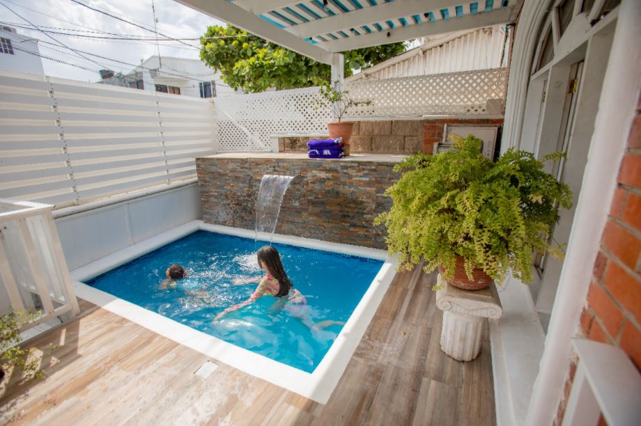 A private, rectangular swimming pool is surrounded by a wooden deck, featuring a small waterfall cascading down a stone wall. Two children are playfully splashing in the clear blue water, while lush greenery is present in a potted plant nearby.
