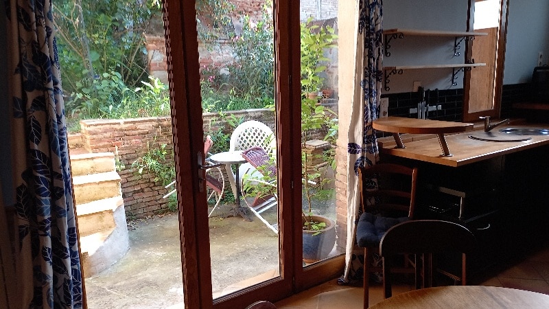 A view of an outdoor space is visible through glass doors. Lush greenery surrounds a small patio with a white chair and a table. Stairs lead to a garden area, while an inviting wooden dining table is seen in the foreground.