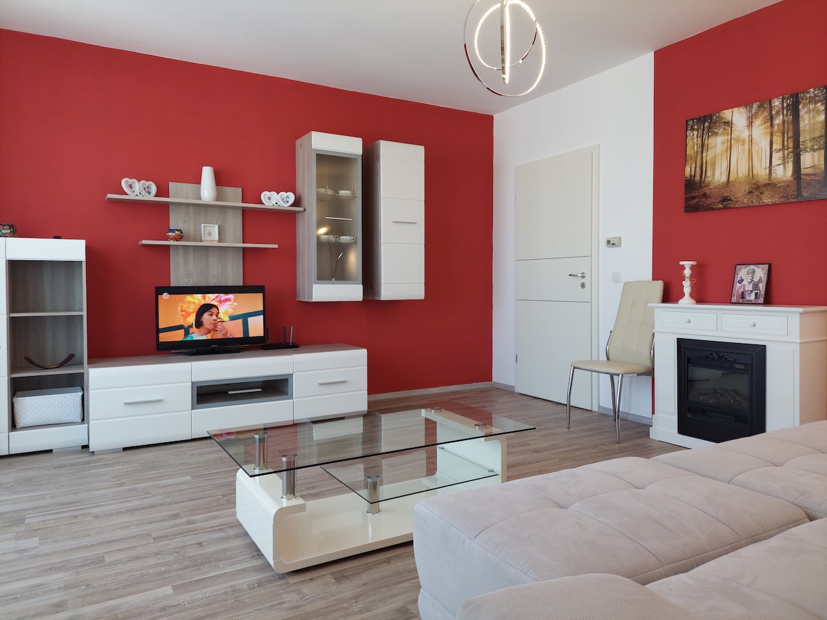 A modern living area features a bright red accent wall and light wood flooring. A sleek glass coffee table sits at the center, complemented by a comfortable sectional sofa. A television is visible on a white entertainment unit, and a decorative electric fireplace adds warmth to the space.