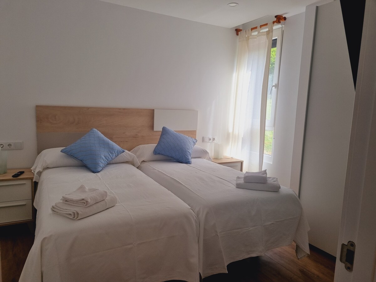 A bright bedroom features two individual beds with blue accent pillows. Light linen covers the beds, while towels are neatly arranged at the foot of each bed. Natural light filters in through a window, complemented by sheer curtains that soften the space.