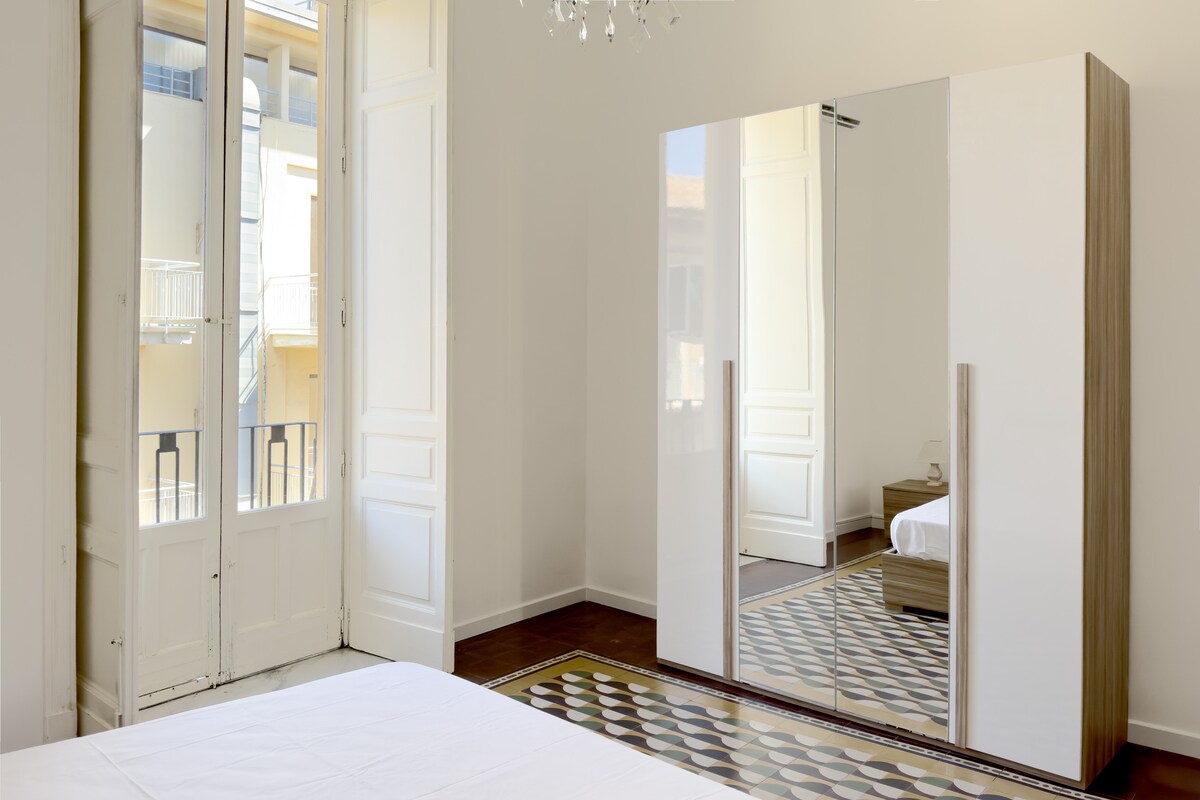 A bright bedroom is showcased, featuring large windows with elegant shutters allowing natural light. A stylish wardrobe with a mirrored front reflects the spacious area. The original patterned floor tiles add character, complementing the minimalist design of the room.