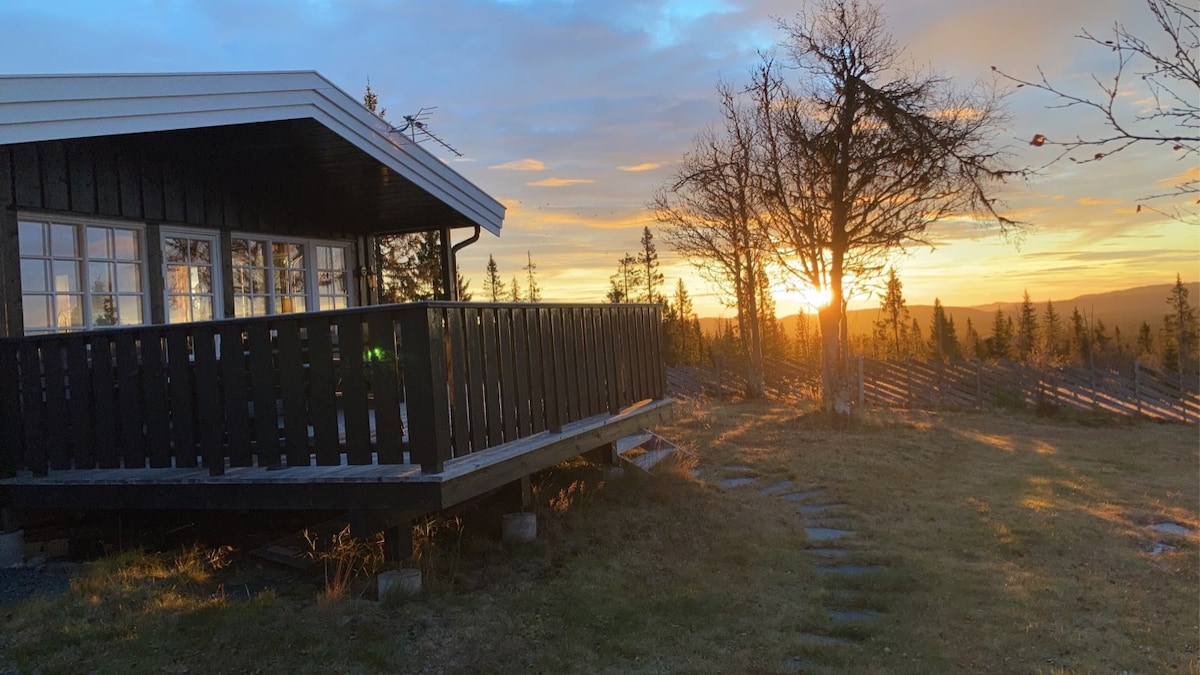 A wooden cabin with large windows is positioned on a hillside, overlooking a sunset that casts warm light. A wooden deck extends from the cabin, and the surrounding landscape features trees and grasses, creating a serene outdoor setting.