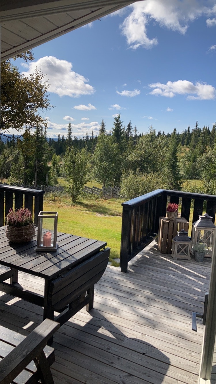A serene outdoor deck features a wooden table and chairs, surrounded by lush greenery and tall trees. Lanterns and potted plants add to the inviting atmosphere. The expansive view reveals a gently sloping landscape under a clear, blue sky with scattered clouds.