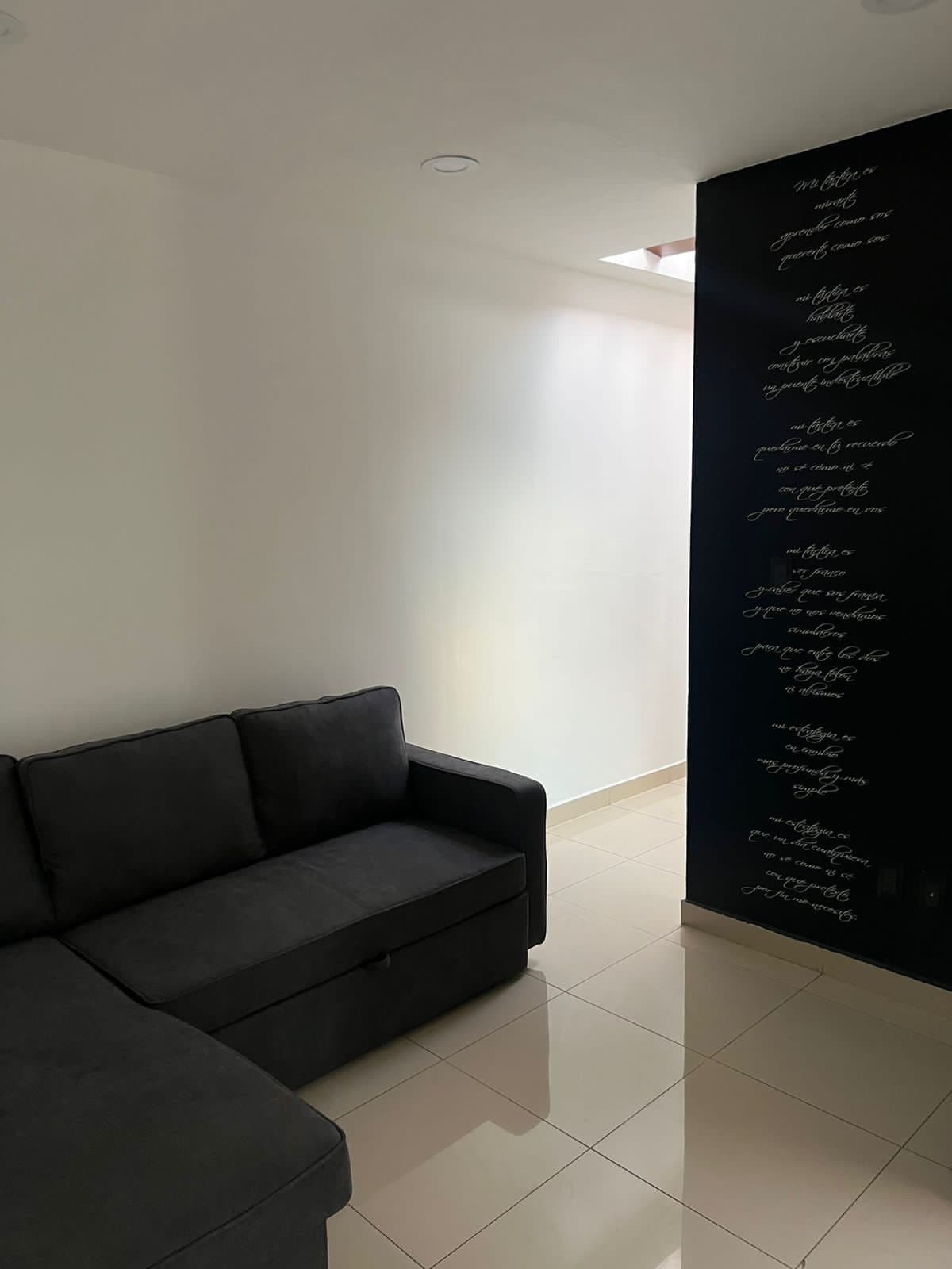 A corner of a cozy living area is shown, featuring a dark sectional couch against a minimalist white wall. Natural light filters in through a skylight, illuminating the space. A contrasting dark wall with unique text adds character to the room.
