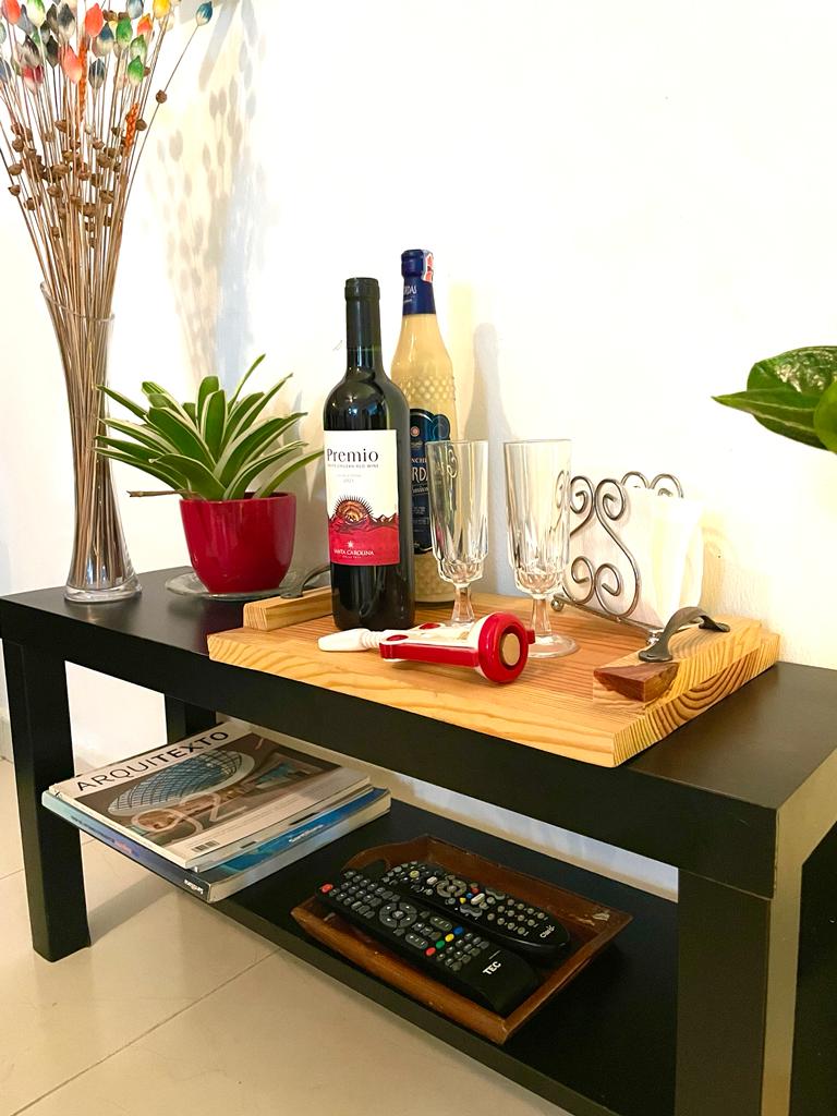 A small black table features a selection of items, including a bottle of red wine and a bottle of a clear beverage. Two elegant glasses are positioned beside a wooden tray that holds a corkscrew and a bottle opener. A potted plant and magazines are situated on a lower shelf.